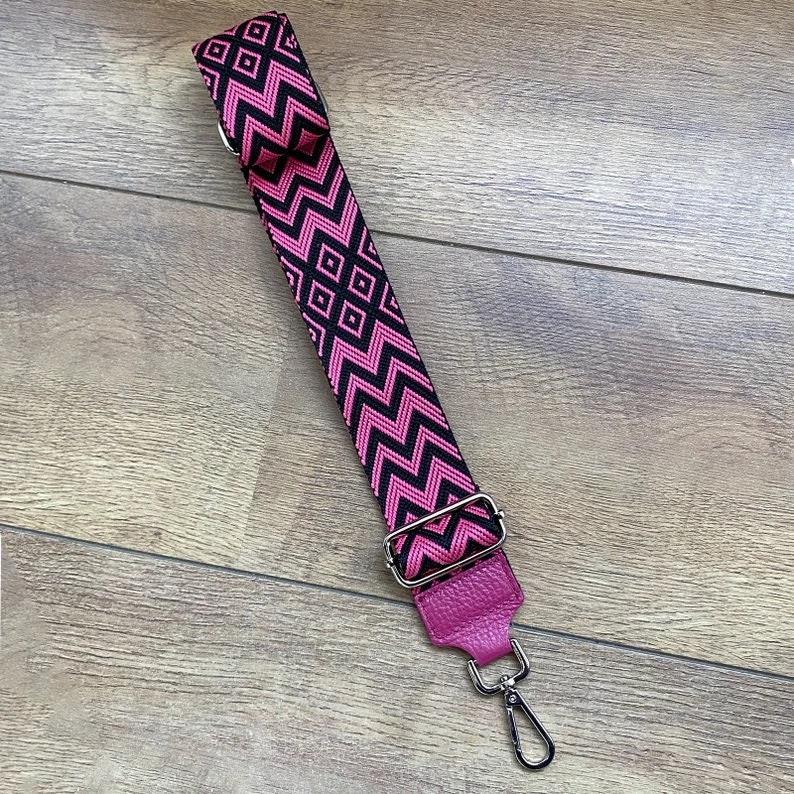 May include: A pink and black patterned adjustable strap with a silver swivel hook. The strap is made of fabric and has a leather end.