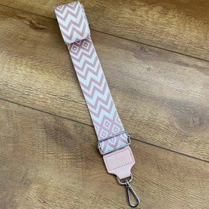 May include: A pink and white patterned strap with a silver metal clasp. The strap is made of woven fabric and has a geometric design.