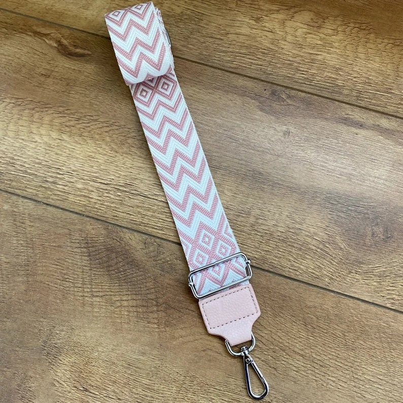 May include: A pink and white patterned strap with a silver metal clasp. The strap is made of woven fabric and has a geometric design.