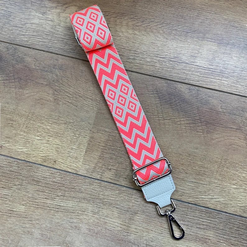 May include: A pink and white patterned adjustable strap with a silver metal clasp. The strap is made of woven fabric and has a leather accent.