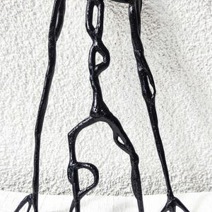 Peut inclure: Une sculpture tridimensionnelle noire et brillante représentant une silhouette humanoïde grande et élancée, dotée de membres allongés, d'une tête arrondie et de mains fines et pointues. La forme présente un aspect organique texturé.