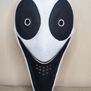 May include: A white mask with a pointed chin and a smooth surface. It features large, oval-shaped black eye patches with small circular cutouts for vision. A wide, black, downward-curving mouth area includes a grid of small ventilation holes.