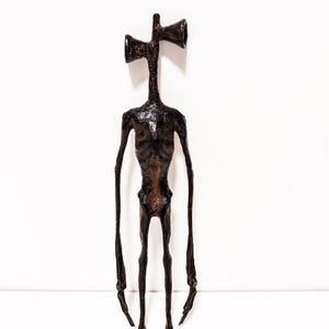 Op de afbeelding: Een donkerbruine Siren Head sculptuur met een lang, slank lichaam en lange ledematen. De kop bestaat uit twee luidsprekerachtige structuren. De figuur staat tegen een witte achtergrond en toont de gedetailleerde textuur van de sculptuur.