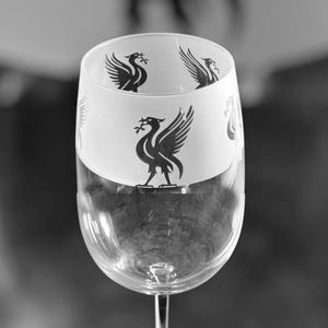 Könnte beinhalten: Ein klares Weinglas mit langem Stiel und rundem Fuß. Der obere Teil des Glases hat ein weißes Band mit einer schwarzen Vogelgrafik. Das Glas ist zum Servieren von Wein bestimmt.