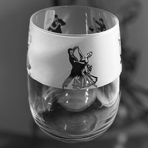 Może przedstawiać: Przezroczysty kieliszek z białym paskiem ozdobionym czarnymi sylwetkami tańczących par. Kieliszek ma szeroki otwór i zaokrągloną podstawę, odpowiedni do napojów. Projekt nawiązuje do tematu tańca towarzyskiego.