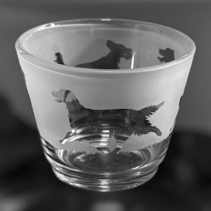 May include: A clear shot glass with a frosted exterior, featuring etched silhouettes of dogs in various poses. The dogs are depicted running through grass. The glass has a slightly flared shape and a thick base, suitable for serving shots.