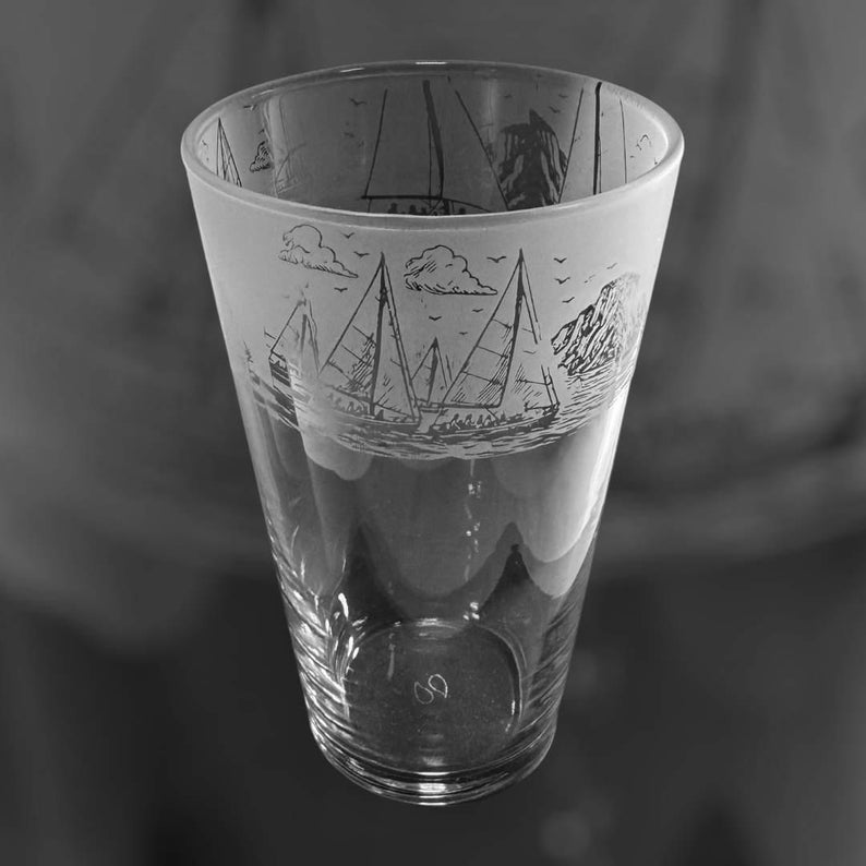 May include: A clear glass tumbler with a nautical design. The upper portion of the glass features a frosted band with etched sailboats, clouds, and birds. The glass has a slightly flared shape and a smooth, transparent finish.