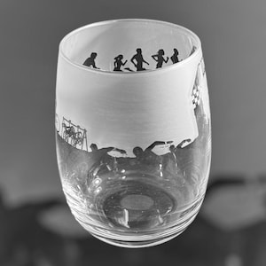 Könnte beinhalten: Ein durchsichtiger Glasbecher mit einem mattierten Design aus Silhouetten von Personen, die laufen und schwimmen. Das Design ist in das Glas geätzt.