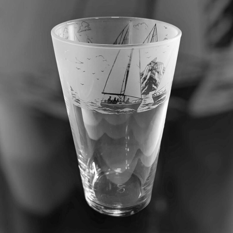 May include: A clear, tall glass with a nautical design. The glass features etched images of sailboats, waves, and a mountain. The top of the glass has a frosted appearance, and the design is in black and white.