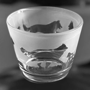 May include: A clear shot glass with a frosted exterior, featuring etched silhouettes of wolves in various poses. The design wraps around the glass, creating a dynamic scene. The glass has a thick base and a slightly flared rim.