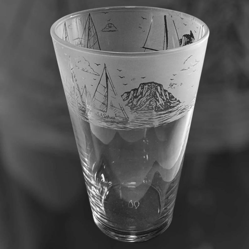 May include: A clear glass with a nautical design. The upper portion of the glass features etched sailboats, clouds, and a mountain scene. The glass is cylindrical with a slightly flared top.