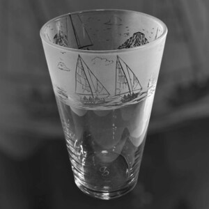 May include: A clear glass with a sailboat design etched around the top. The design features sailboats, mountains, and birds. The glass has a wide mouth and tapers down to a narrower base. The bottom of the glass has a small, etched design.