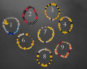 May include: Nine beaded bracelets in various designs are displayed against a dark background. The bracelets feature a variety of beads in colors like yellow, black, red, white, and blue. Some bracelets have letter beads spelling out words.
