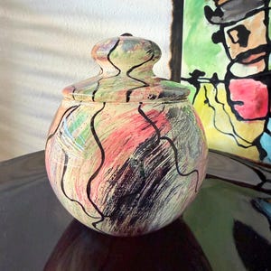 May include: A decorative ceramic jar with a lid. The jar is painted with a colorful abstract design featuring pink, green, and black brushstrokes. The jar has a rounded body and a bulbous lid.