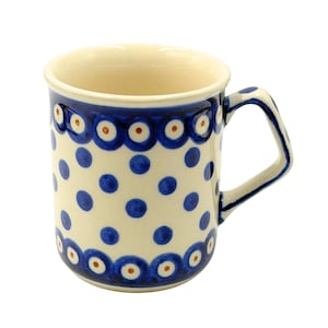 May include: A ceramic mug with a cream-colored base featuring a pattern of blue polka dots and bands of blue and orange circular motifs at the top and bottom. The mug has a single handle on the right side and a smooth, glossy finish throughout.