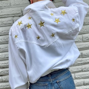 May include: White jacket with gold star embroidery on the back. The jacket has a high collar and a cinched waist. The model is wearing blue jeans. The stars are of varying sizes and are evenly spaced across the back of the jacket.