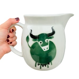May include: White ceramic pitcher with a green bull illustration. The bull has black horns and a white face. The pitcher has a curved handle and a spout for pouring. The pitcher is held by a hand with dark red nail polish.