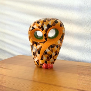 May include: A decorative owl figurine with a mosaic design. The owl features a vibrant orange body with brown, tan, and black mosaic tiles. It has large, round eye openings with green accents and small red feet. The figurine is on a wooden surface.