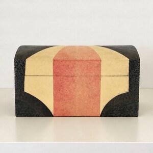 May include: A rectangular box with a curved lid featuring a geometric pattern. The surface has a textured finish in black, cream, and coral pink. The design consists of a central coral vertical stripe flanked by cream panels and black edges.