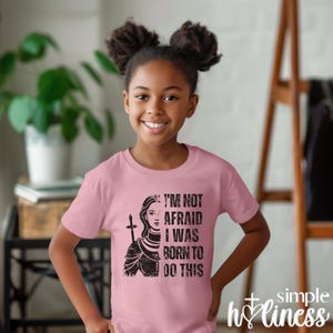 May include: A light pink t-shirt featuring a graphic of a woman warrior with the text "I'M NOT AFRAID I WAS BORN TO DO THIS." The shirt also has the words "simple holiness" in the bottom right corner.