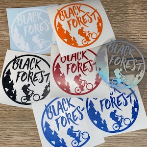 May include: A collection of circular stickers with the text "BLACK FOREST" in various colors. Each sticker features a silhouette of a person riding a mountain bike, with trees in the background. Colors include black, orange, red, blue, and iridescent.