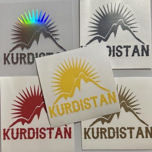 Op de afbeelding: Vijf vinyl stickers met een bergketen en zon ontwerp. De stickers zijn in verschillende kleuren: goud, rood, zwart, bruin en iriserend. Elke sticker heeft het woord "Kurdistan" onder het ontwerp gedrukt.