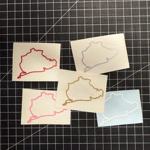 以下が含まれることがあります： 異なる色のニュルブルクリンク・レースコースのステッカー5枚。赤、銀、金、ピンク、白。ステッカーは黒いグリッドの背景に貼られています。