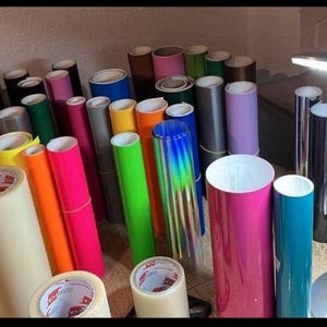 May include: A variety of colored vinyl rolls, including red, orange, yellow, green, blue, purple, black, white, and silver. The rolls are arranged in a row on a table, with a few rolls stacked on top of each other. There is a white label on each roll with black text.