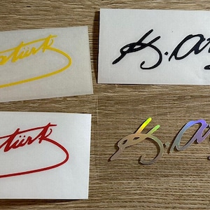 Könnte beinhalten: Vier verschiedene Versionen der Unterschrift von Mustafa Kemal Atatürk auf weißem Papier. Die Unterschriften sind in Schwarz, Gelb, Rot und schillernd.