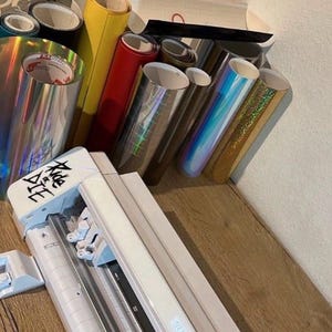 May include: A white vinyl cutter with a roll of red vinyl and a variety of iridescent vinyl rolls. The vinyl cutter has a black and white label that says "Silhouette".