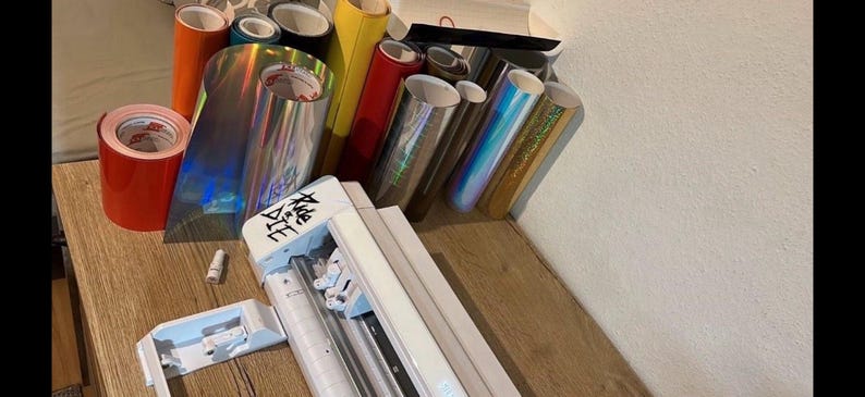 May include: A white vinyl cutter with a roll of red vinyl and a variety of iridescent vinyl rolls. The vinyl cutter has a black and white label that says "Silhouette".