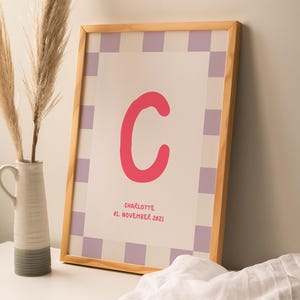 Könnte beinhalten: Ein gerahmter Druck in einem hellen Holzrahmen mit einem Schachbrettmuster-Rand in Weiß und Helllila. In der Mitte befindet sich ein großer rosa Buchstabe C, darunter der Text "CHARLOTTE" und "01. NOVEMBER 2021" in roter Schrift.