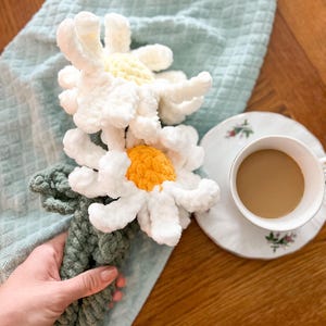 May include: Two crocheted white daisies with yellow centers are tied together with a green rope. The daisies are on a light blue towel, and a cup of coffee is in the background.