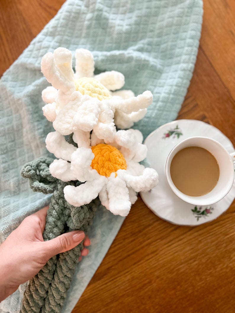 May include: Two crocheted white daisies with yellow centers are tied together with a green rope. The daisies are on a light blue towel, and a cup of coffee is in the background.
