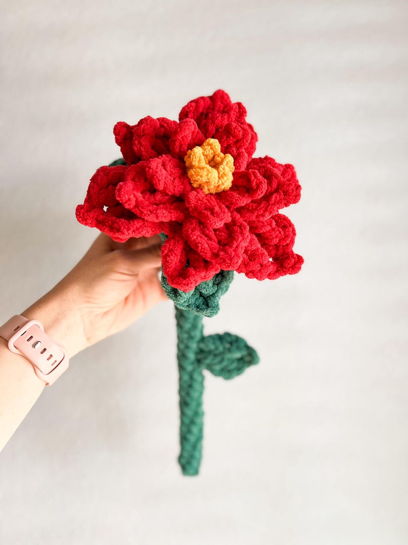 May include: A crocheted red poinsettia flower with a yellow center and green stem and leaves.