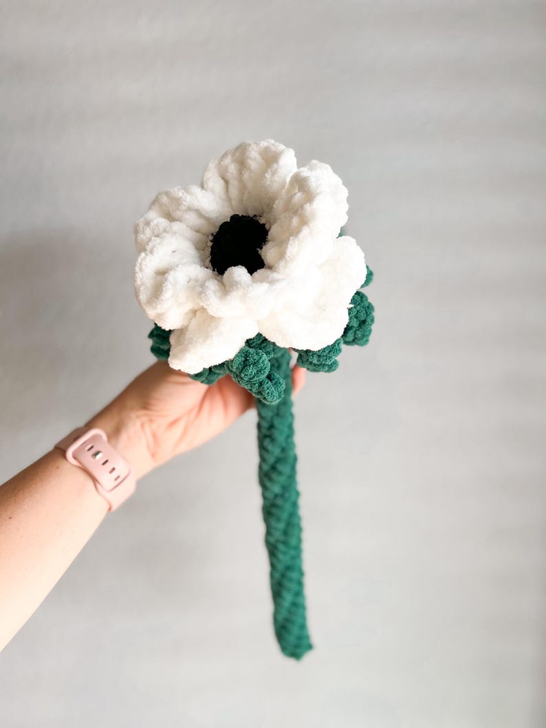 May include: A white crocheted flower with a black center and a green crocheted stem.
