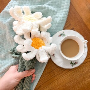 May include: A crocheted bouquet of white daisies with yellow centers, held in a hand. The bouquet is on a light blue towel, next to a cup of coffee on a white saucer.