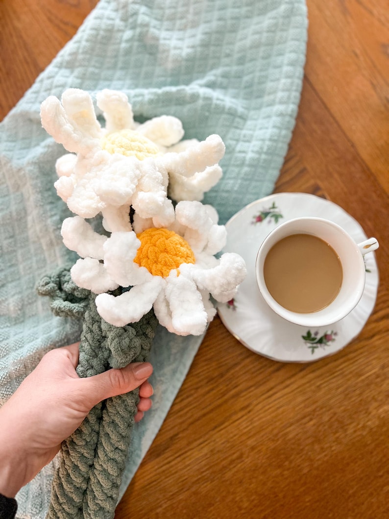 May include: A crocheted bouquet of white daisies with yellow centers, held in a hand. The bouquet is on a light blue towel, next to a cup of coffee on a white saucer.