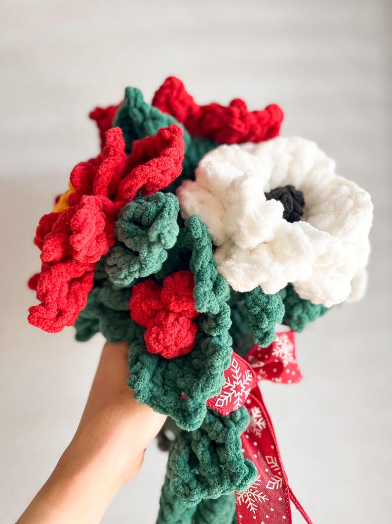 May include: A crocheted bouquet of red poinsettia flowers, white flowers, and green leaves tied with a red ribbon with white snowflakes.