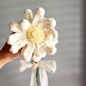 May include: A white crocheted daisy with a yellow center, tied with a white ribbon, and a green stem.