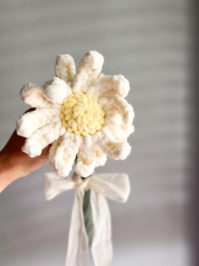 May include: A white crocheted daisy with a yellow center, tied with a white ribbon, and a green stem.