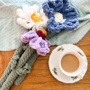 May include: A bouquet of crocheted flowers in shades of white, blue, purple, and green. The flowers are arranged on a wooden table with a cup of coffee and a saucer.