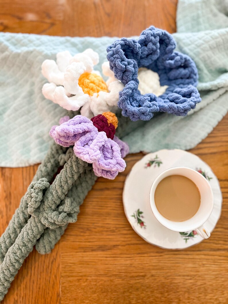 May include: A bouquet of crocheted flowers in shades of white, blue, purple, and green. The flowers are arranged on a wooden table with a cup of coffee and a saucer.