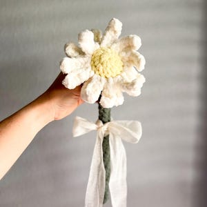 May include: A crocheted daisy flower with a green stem and a white ribbon. The flower is white with a yellow center.