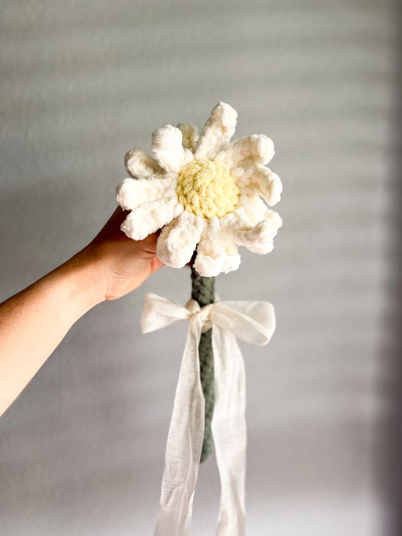 May include: A crocheted daisy flower with a green stem and a white ribbon. The flower is white with a yellow center.