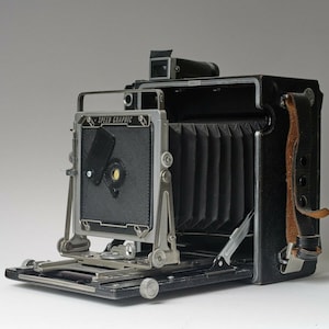 Peut inclure: Un appareil photo Speed Graphic noir avec un soufflet et une sangle en cuir. L'appareil photo a un dos en métal argenté et un bouton de mise au point en métal argenté.