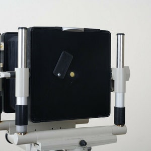 May include: A large format camera with a black bellows and a silver and black body. The camera is mounted on a tripod.