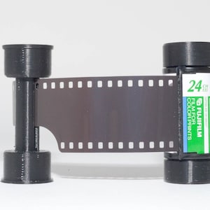 May include: A black and green 35mm film canister with the text "FUJIFILM FILM FOR COLOR PRINTS" and "24" printed on the side. The film is partially exposed and shows a black and white image.