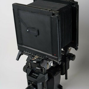 Peut inclure: Un appareil photo grand format noir avec un soufflet et une marque Sinar sur la base. L'appareil est monté sur un trépied.