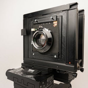 May include: A black large format camera with a lens attached. The camera has a black leather bellows and a metal body. The camera is mounted on a tripod.